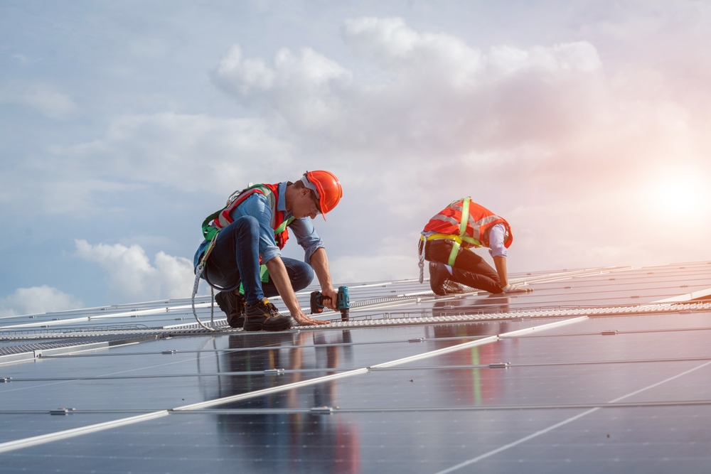 
Technician wearing safety harness belt using drill during installing the solar panels on roof structure of building factory.